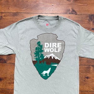 May include: A light green t-shirt with a graphic of a white dire wolf in a green and brown national park emblem. The emblem says "Dire Wolf" in white text.