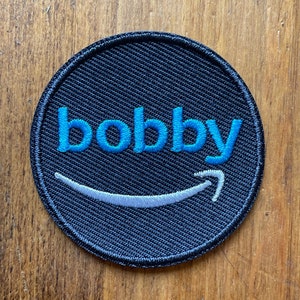 May include: A black circular patch with a blue embroidered word "bobby" and a white embroidered Amazon smile.