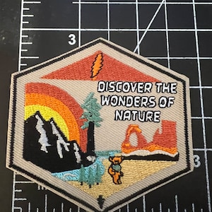 May include: A hexagonal embroidered patch with a beige background. The patch features a sunset scene with mountains, a tree, a river, and a bear. The text "DISCOVER THE WONDERS OF NATURE" is printed in white on the patch.