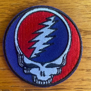 May include: A circular embroidered patch featuring a stylized skull design with a lightning bolt across the forehead. The design uses a vibrant red and blue background with a white skull outline and a white lightning bolt, all bordered by a dark blue edge.
