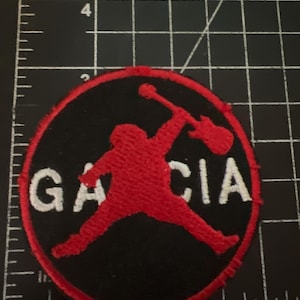 May include: A circular embroidered patch featuring a red silhouette of a person holding a guitar above the text GARCIA in white, set against a black background with a red border, positioned on a black cutting mat with white grid lines.