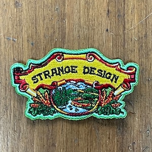 May include: A green, yellow, and red embroidered patch with the text "Strange Design" in a banner. The patch features a green, blue, and red landscape scene with a river and mountains.