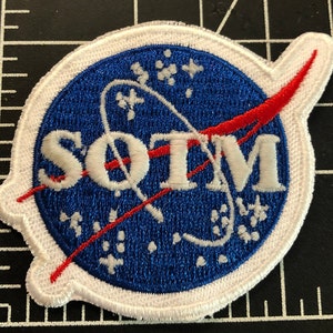 May include: A white embroidered patch with a blue circle and a red curved line. The circle has white stars and the text "SOTM" in white.