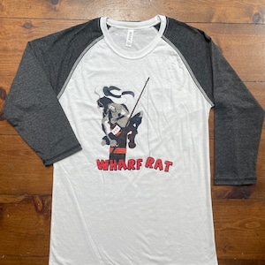 May include: White and charcoal raglan baseball tee with a graphic of a cartoon rat fishing. The rat wears a hat and holds a fishing rod. The text "WHARF RAT" is printed in red below the graphic.