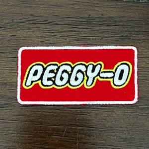 May include: A rectangular red patch with the text "PEGGY-O" in white, outlined in yellow. The patch has a white border and is likely an iron-on or sew-on patch for clothing or accessories.
