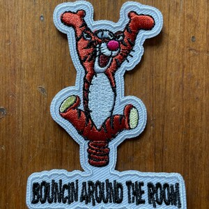 May include: An embroidered iron-on patch featuring a cartoon tiger with orange and black stripes, a pink nose, and a white background. The tiger is jumping up and down with its legs in the air. The text "BOUNCIN' AROUND THE ROOM" is written in black below the tiger.