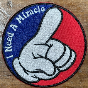May include: A round, red and blue embroidered patch with a white cartoon hand pointing to the right. The text "I Need A Miracle" is written in white on the blue portion of the patch.