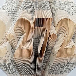 Puede incluir: Una escultura de arte de libro hecha de p&aacute;ginas dobladas de un libro, formando el n&uacute;mero 2021. La escultura est&aacute; hecha de papel color crema y tiene un efecto tridimensional.