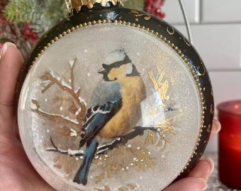 Op de afbeelding: Een ronde kerstbal met een gedetailleerde schildering van een blauw-gele vogel op een tak. De ornament heeft een zwarte rand met gouden accenten en een gouden dop. De achtergrond is een besneeuwde scène.