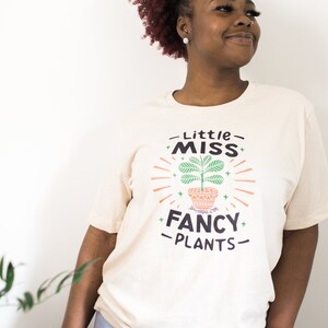 May include: A woman wearing a cream colored t-shirt with the text "Little Miss Fancy Plants" and a graphic of a potted plant with green leaves.