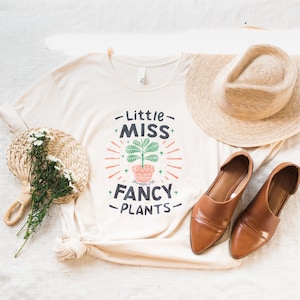 May include: A white t-shirt with the text "Little Miss Fancy Plants" and a stylized illustration of a potted plant. The shirt is laid out on a white surface with a straw hat, a pair of brown shoes, and a woven fan with white flowers.