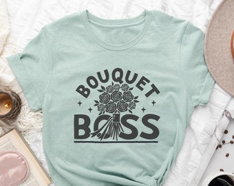 May include: A light blue t-shirt with the text "BOUQUET BOSS" in black lettering. The text is surrounded by a stylised bouquet of roses.