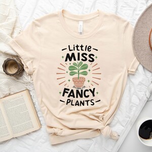 May include: A beige t-shirt with a graphic design of a potted plant and the text "Little Miss Fancy Plants".