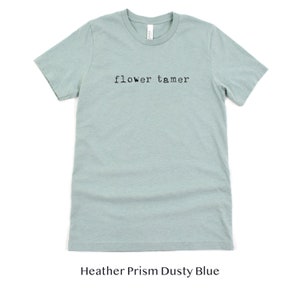 May include: A light blue t-shirt with the text "flower tamer" printed in black on the front. The shirt is made of a soft, comfortable material and is a heather prism dusty blue color.