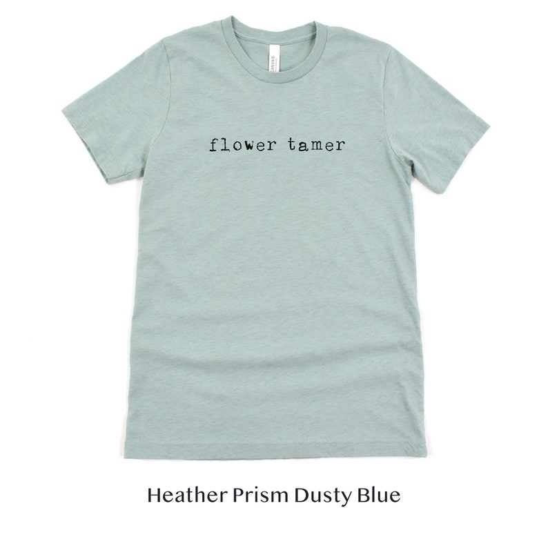 May include: A light blue t-shirt with the text "flower tamer" printed in black on the front. The shirt is made of a soft, comfortable material and is a heather prism dusty blue color.