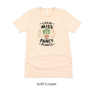 May include: A soft cream colored t-shirt with a graphic design that says "Little Miss Fancy Plants" with a potted plant illustration.