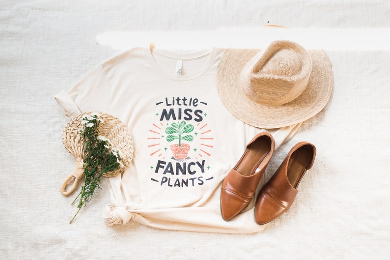 May include: A white t-shirt with the text "Little Miss Fancy Plants" and a stylized illustration of a potted plant. The shirt is laid out on a white surface with a straw hat, a pair of brown shoes, and a woven fan with white flowers.