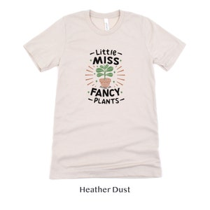 May include: A heather dust colored t-shirt with a graphic of a potted plant and the text "Little Miss Fancy Plants".