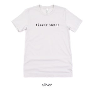 May include: A silver t-shirt with the text "flower tamer" printed in black.
