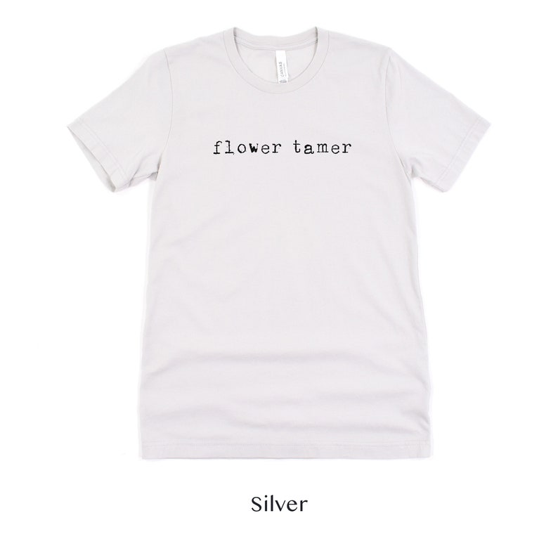 May include: A silver t-shirt with the text "flower tamer" printed in black.