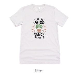 May include: A white t-shirt with a graphic design of a potted plant and the text "Little Miss Fancy Plants".