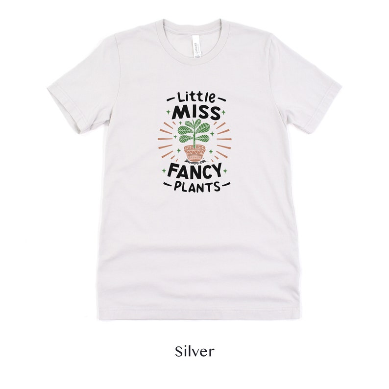 May include: A white t-shirt with a graphic design of a potted plant and the text "Little Miss Fancy Plants".