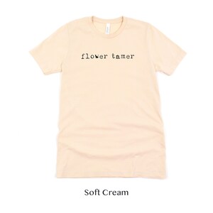May include: A soft cream colored t-shirt with the text "flower tamer" printed in black on the front.