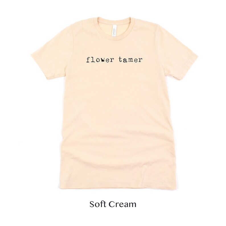 May include: A soft cream colored t-shirt with the text "flower tamer" printed in black on the front.