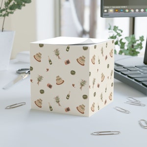 May include: A white square notepad with a repeating pattern of wedding cake, champagne bottles, flowers, and cameras. The notepad is on a white desk with paper clips.