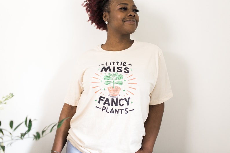 May include: A woman wearing a cream colored t-shirt with the text "Little Miss Fancy Plants" and a graphic of a potted plant with green leaves.