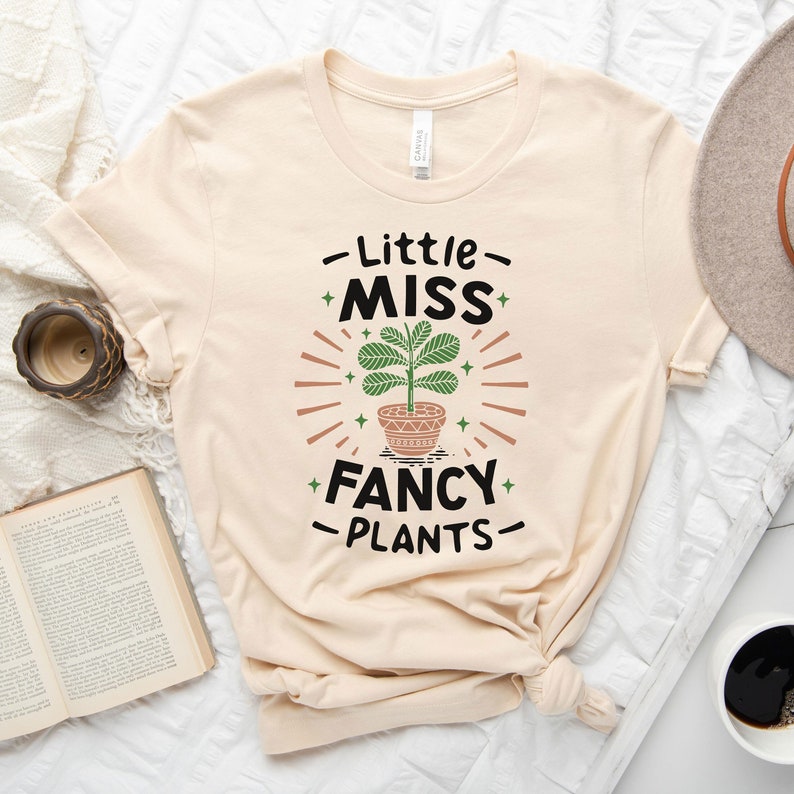 May include: A beige t-shirt with a graphic design of a potted plant and the text "Little Miss Fancy Plants".