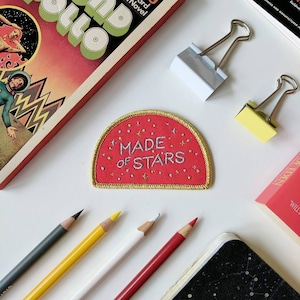 May include: A red fabric patch with gold stitching and a white embroidered message that reads "MADE OF STARS". The patch is shaped like a half-circle and has small gold stars scattered around the edges.