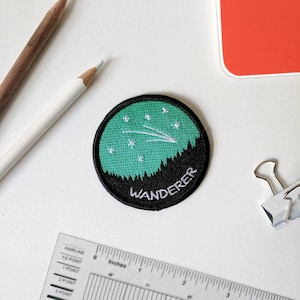 May include: A green and black embroidered patch with a white shooting star and the word "WANDERER" in white text.