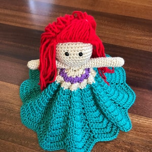 May include: A handmade crochet doll with red yarn hair, black button eyes, and a cream-colored face. The doll wears a teal dress with a scalloped edge and a purple and white necklace. The doll is lying on a wooden surface.