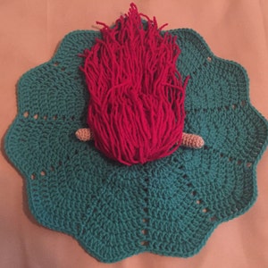 May include: A handmade crochet piece featuring a teal scalloped base with a central design of bright pink yarn resembling flames. Two small, beige crochet pieces are positioned near the base of the pink yarn. The piece is likely a decorative item or a unique craft.