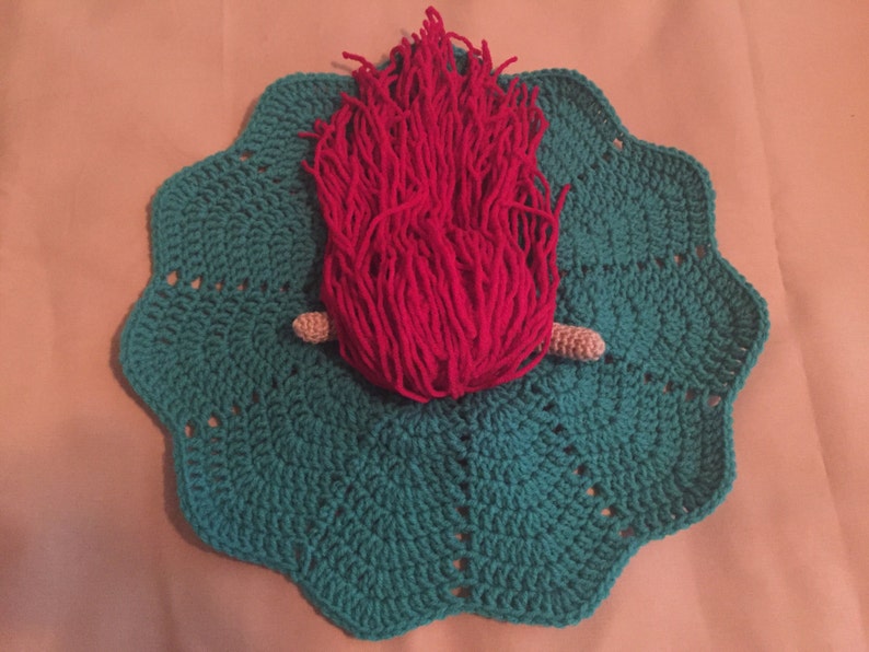 May include: A handmade crochet piece featuring a teal scalloped base with a central design of bright pink yarn resembling flames. Two small, beige crochet pieces are positioned near the base of the pink yarn. The piece is likely a decorative item or a unique craft.