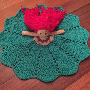May include: A handmade crochet doll with red yarn hair and a teal blanket. The doll has a beige body, a purple and beige collar, and black eyes. The blanket is scalloped around the edges.