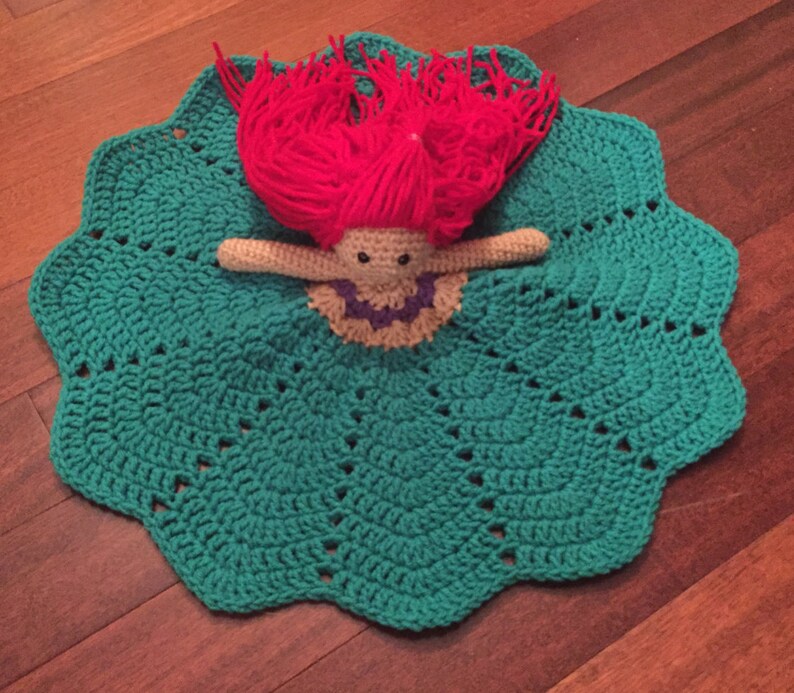May include: A handmade crochet doll with red yarn hair and a teal blanket. The doll has a beige body, a purple and beige collar, and black eyes. The blanket is scalloped around the edges.