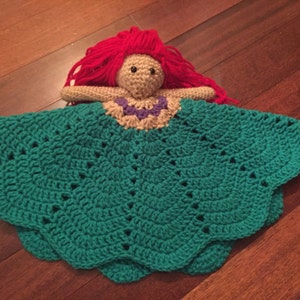 May include: A handmade crochet doll with red yarn hair, a tan face, and a teal dress. The doll's dress has a scalloped edge and a decorative neckline. The doll is lying on a wooden surface.