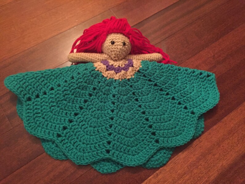 May include: A handmade crochet doll with red yarn hair, a tan face, and a teal dress. The doll's dress has a scalloped edge and a decorative neckline. The doll is lying on a wooden surface.