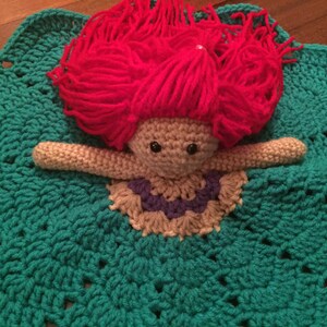 May include: A handmade crochet doll with red yarn hair, beige arms, and a purple and beige dress, resting on a teal crochet blanket. The blanket has a scalloped edge and a textured pattern, creating a whimsical and cozy feel.