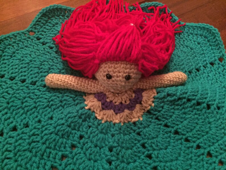 May include: A handmade crochet doll with red yarn hair, beige arms, and a purple and beige dress, resting on a teal crochet blanket. The blanket has a scalloped edge and a textured pattern, creating a whimsical and cozy feel.