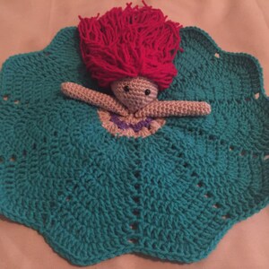 May include: A handmade crochet doll with a teal dress and a scalloped edge. The doll has a pink yarn hair and a beige body. The doll's dress has a purple and white neckline.