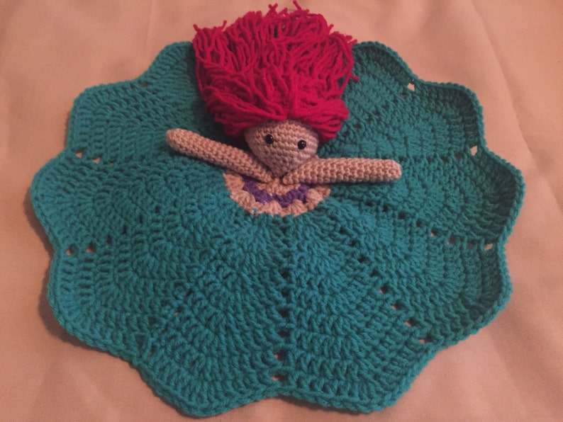 May include: A handmade crochet doll with a teal dress and a scalloped edge. The doll has a pink yarn hair and a beige body. The doll's dress has a purple and white neckline.