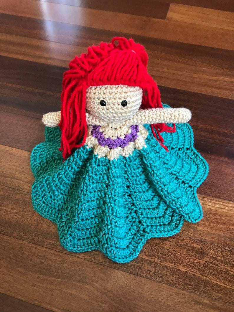 May include: A handmade crochet doll with red yarn hair, black button eyes, and a cream-colored face. The doll wears a teal dress with a scalloped edge and a purple and white necklace. The doll is lying on a wooden surface.