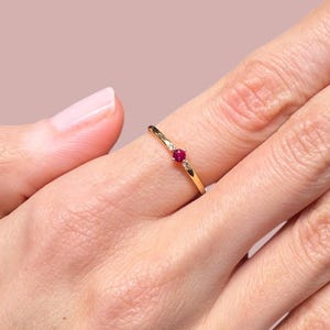 May include: A delicate gold ring featuring a single ruby gemstone. The ring is thin and has a simple design.