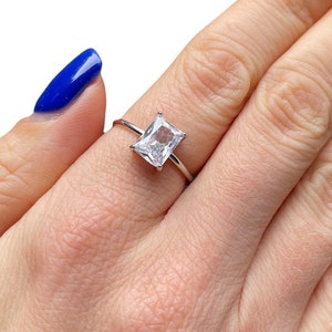 May include: A silver ring with a clear square-shaped gemstone. The ring is on a person's finger.