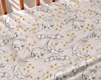 May include: A white fabric sheet featuring a repeating pattern of black line-drawn crescent moons with faces, circular sun-like faces, and small mustard yellow stars. Each moon is accompanied by a banner containing the text la luna.