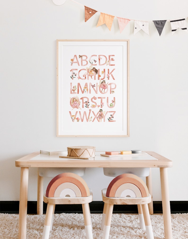 May include: A framed alphabet poster featuring pink letters and illustrations of fairies and flowers. The poster is hanging on a white wall above a wooden table with two rainbow-shaped chairs.