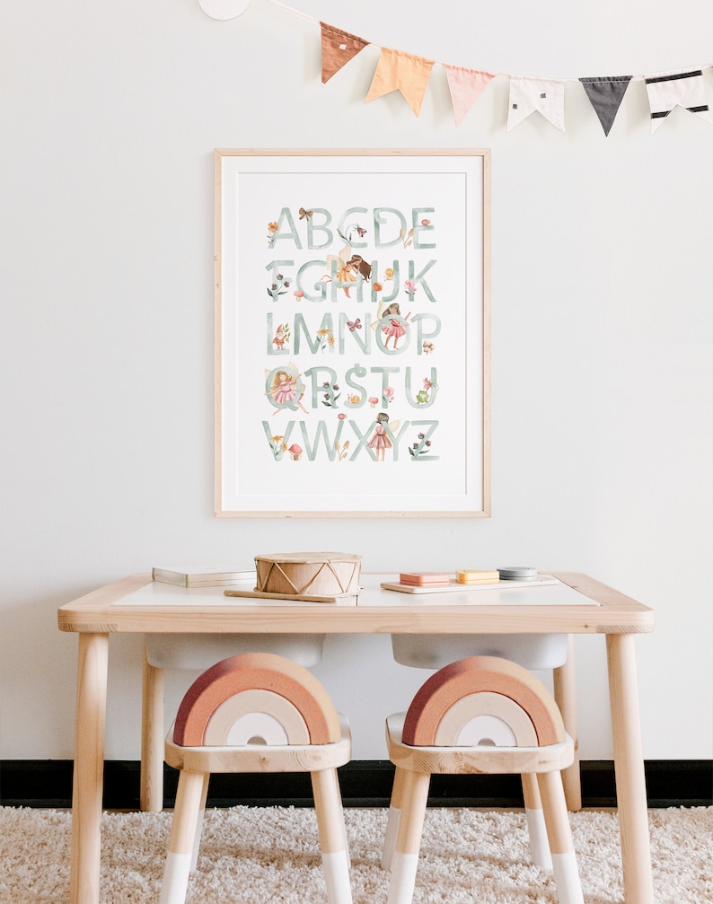 May include: A framed alphabet poster with a green background and illustrations of fairies and flowers. The poster is hanging on a white wall above a wooden table with two rainbow-shaped chairs.
